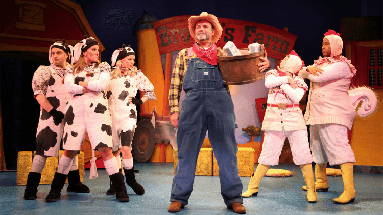 Image of performers on stage. A farmer stand in the middle with actors dressed as chickens and cows on either side of him